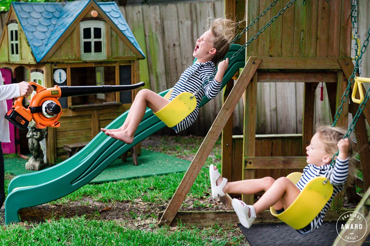 Two children in matching yellow outfits enjoy swinging together on a backyard playset with a green slide and playhouse.