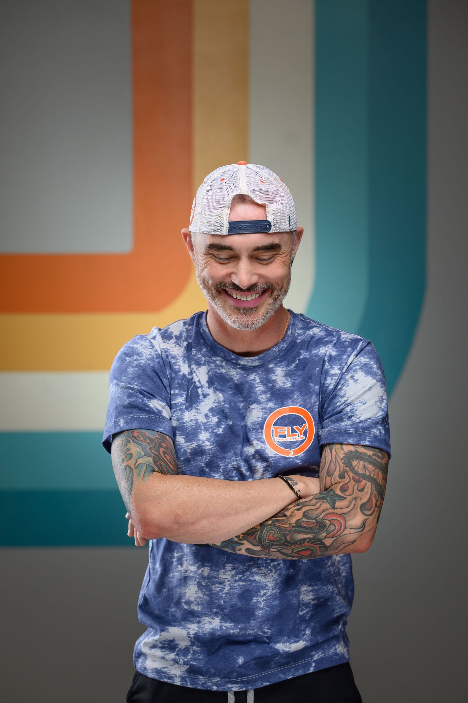Person in tie-dye shirt and baseball cap poses playfully against geometric wall design.