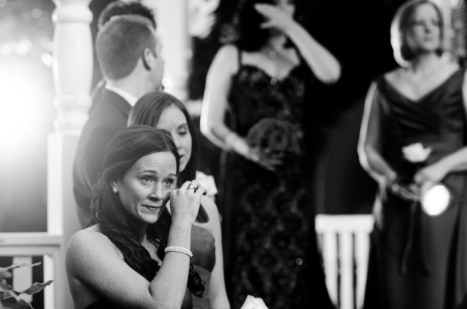 Black and white wedding photo of emotional guest wiping tears during ceremony, with other guests in elegant attire in background.