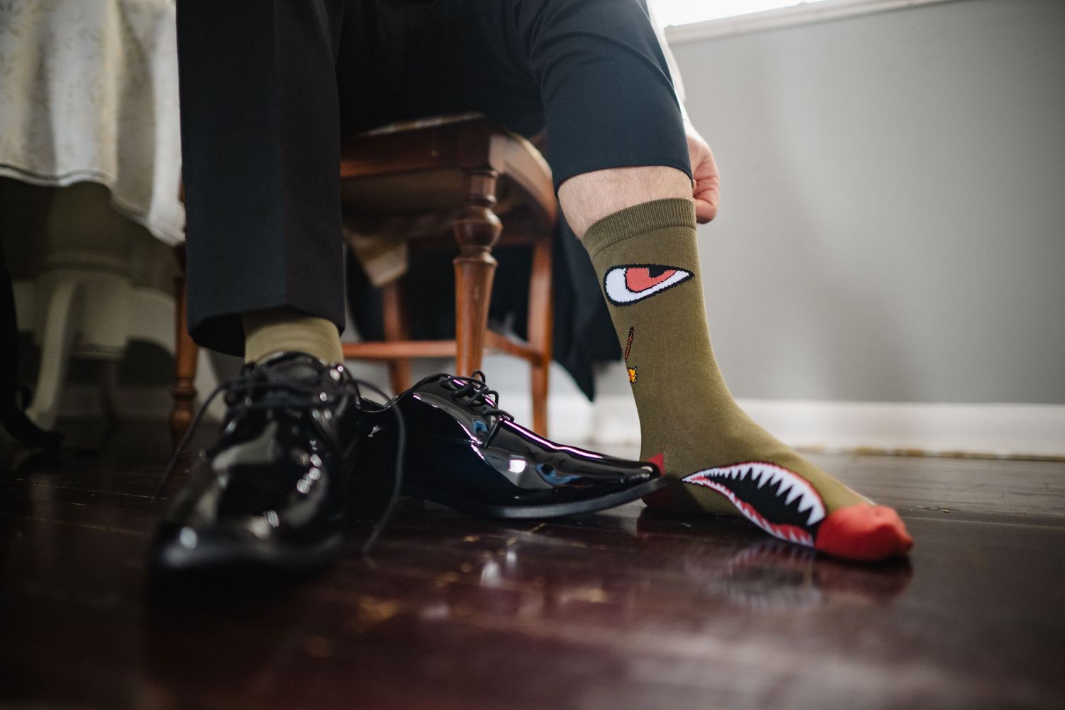 Man putting on olive green shark design novelty socks next to shiny black dress shoes on hardwood floor.