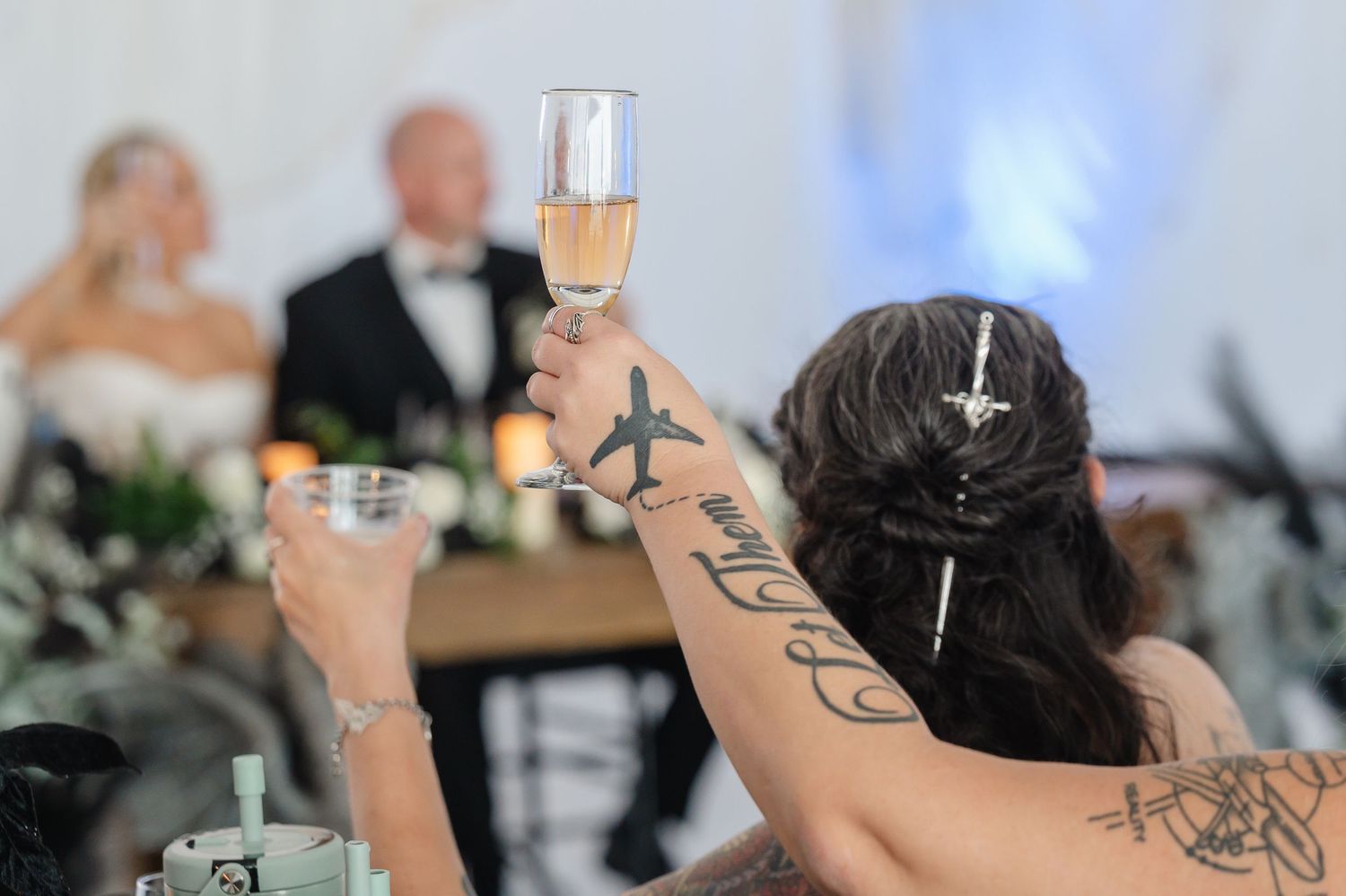 Wedding guest raises champagne toast showing airplane tattoo on arm during reception celebration.