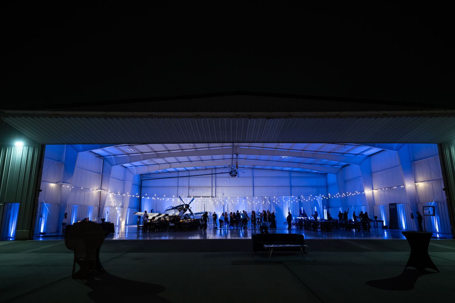 A grand piano performance illuminated by blue lights inside an open aircraft hangar at night.