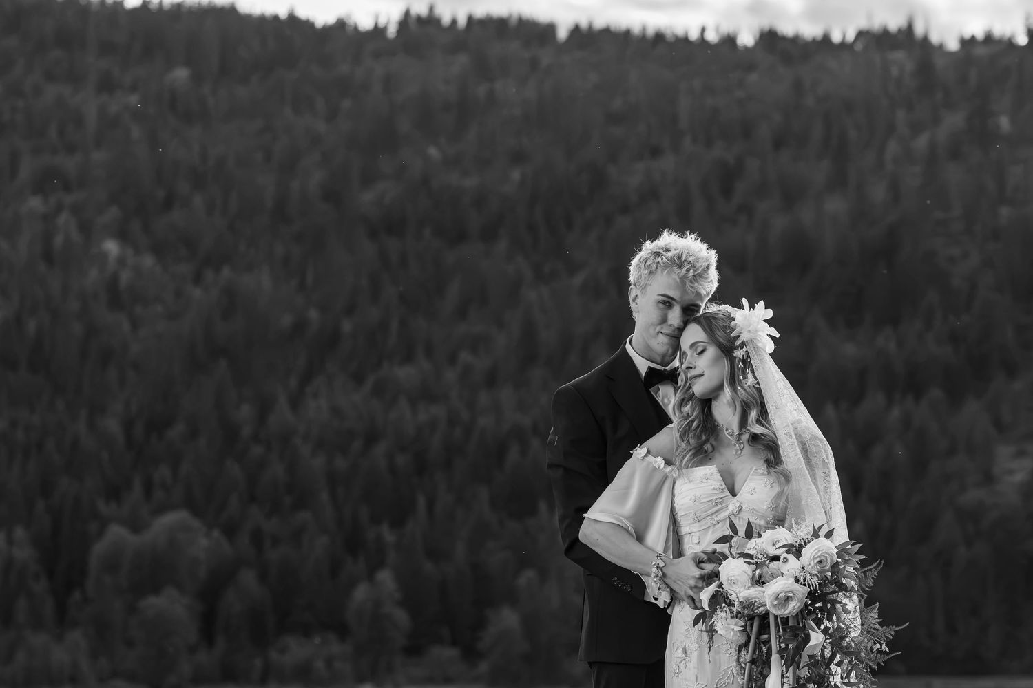 Black and white photo of a bride and groom embracing with a lush forest backdrop on their wedding day.