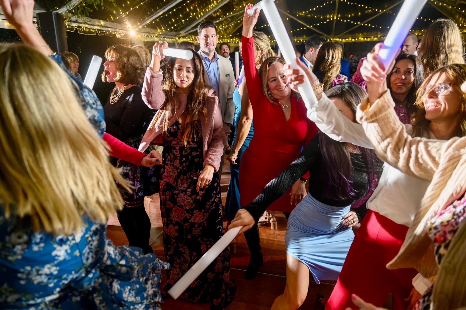 Energetic crowd dances with glowing foam sticks on a dance floor under string lights.