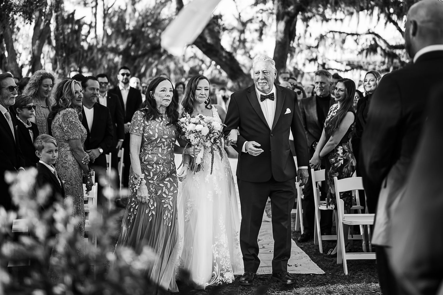 Black and white photo of bridal party walking down outdoor aisle with Spanish moss draped trees in background.
