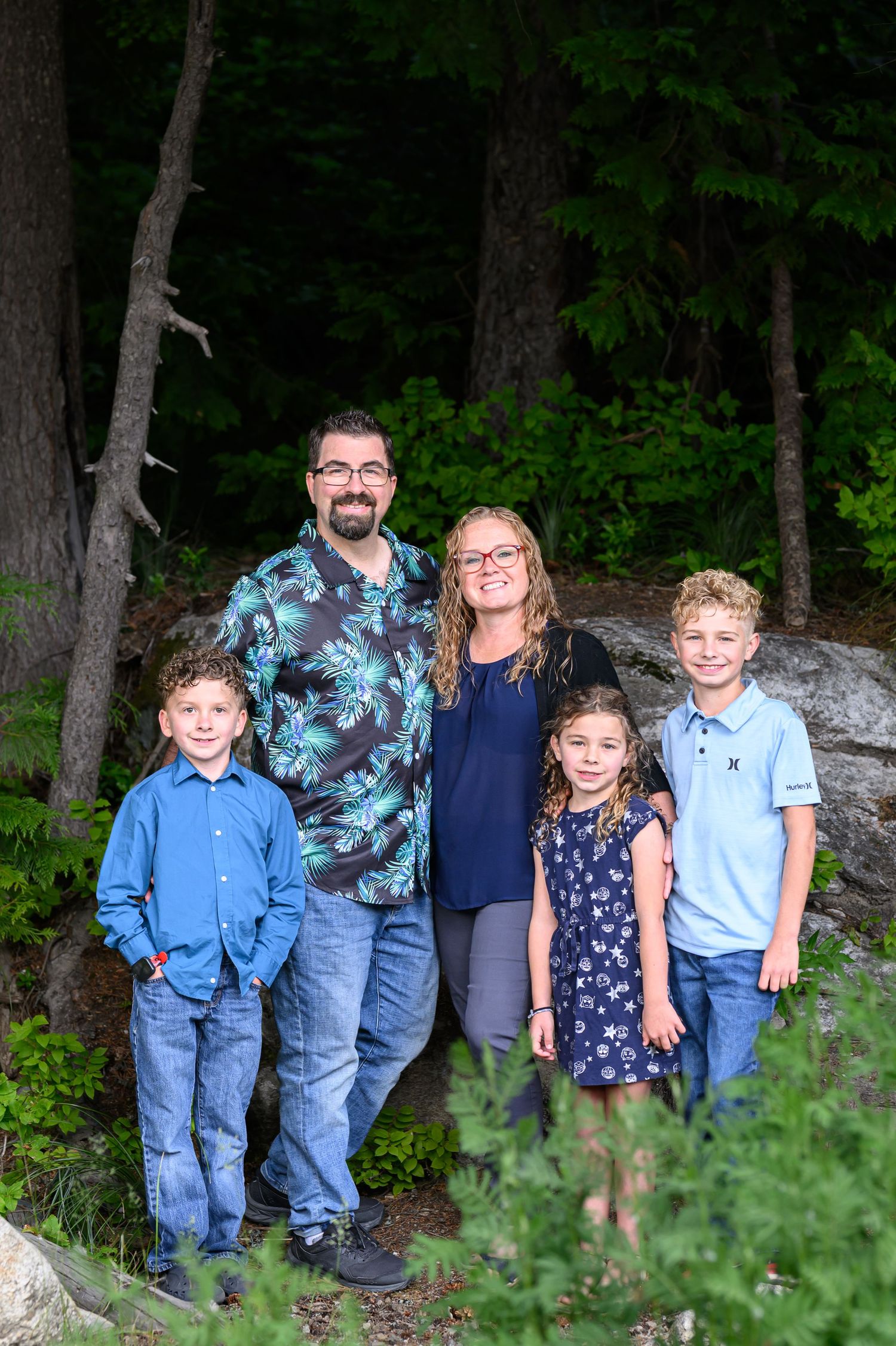 Family of five smiling together outdoors in a lush forest setting, parents with three children in casual attire.