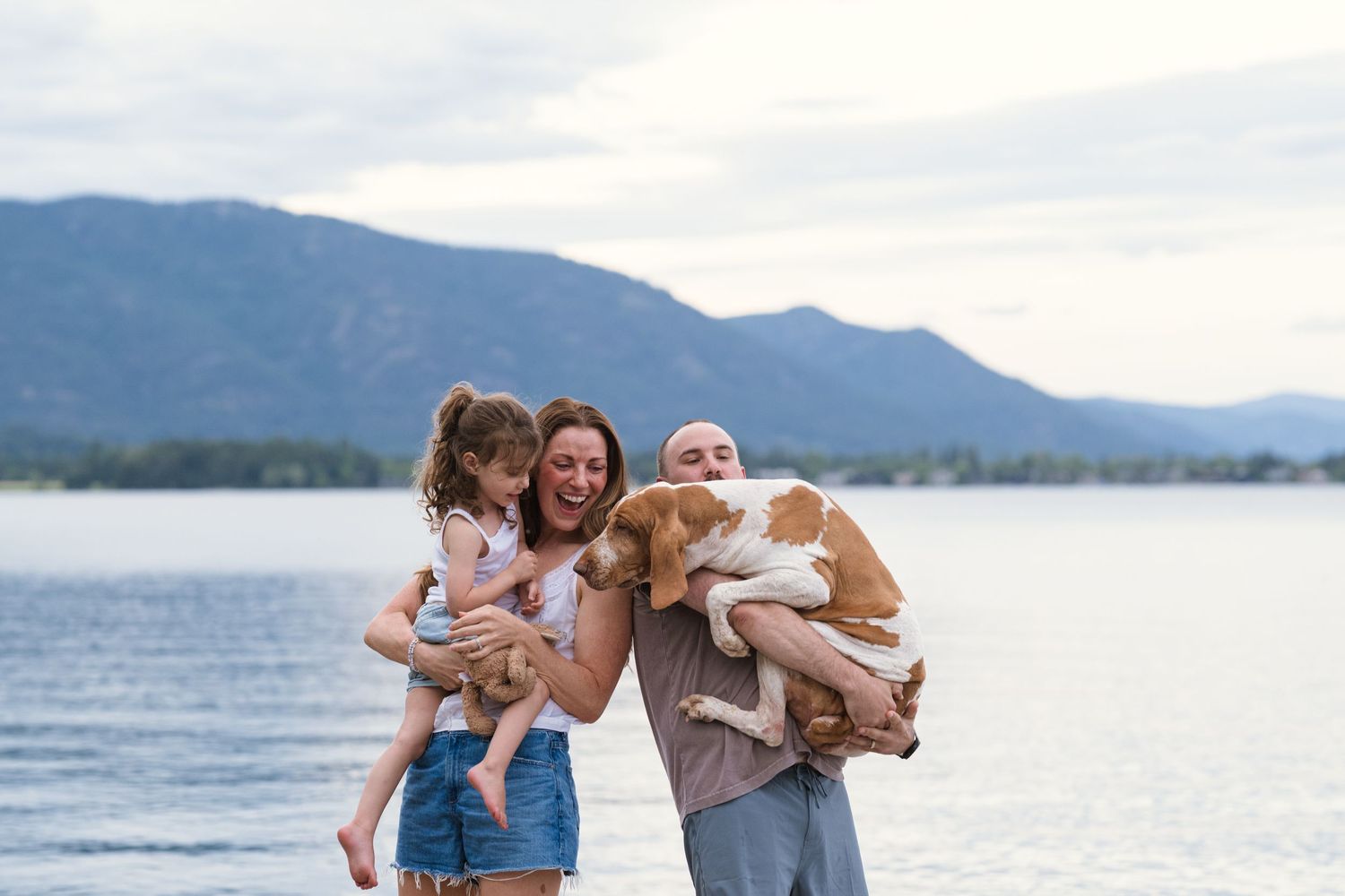 Happy family with young girl and dog posing by a mountain lake on a cloudy day.