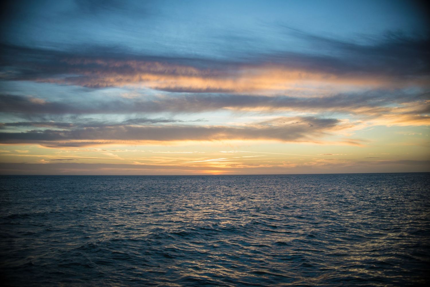Dramatic sunset over calm ocean waters with vibrant orange and pink clouds reflecting on the dark blue sea surface.