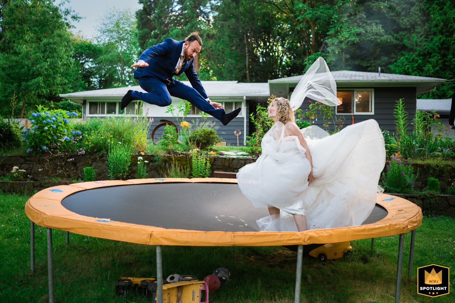 Playful bride and groom jumping together on a backyard trampoline, one wearing a flowing wedding dress at their wedding reception.