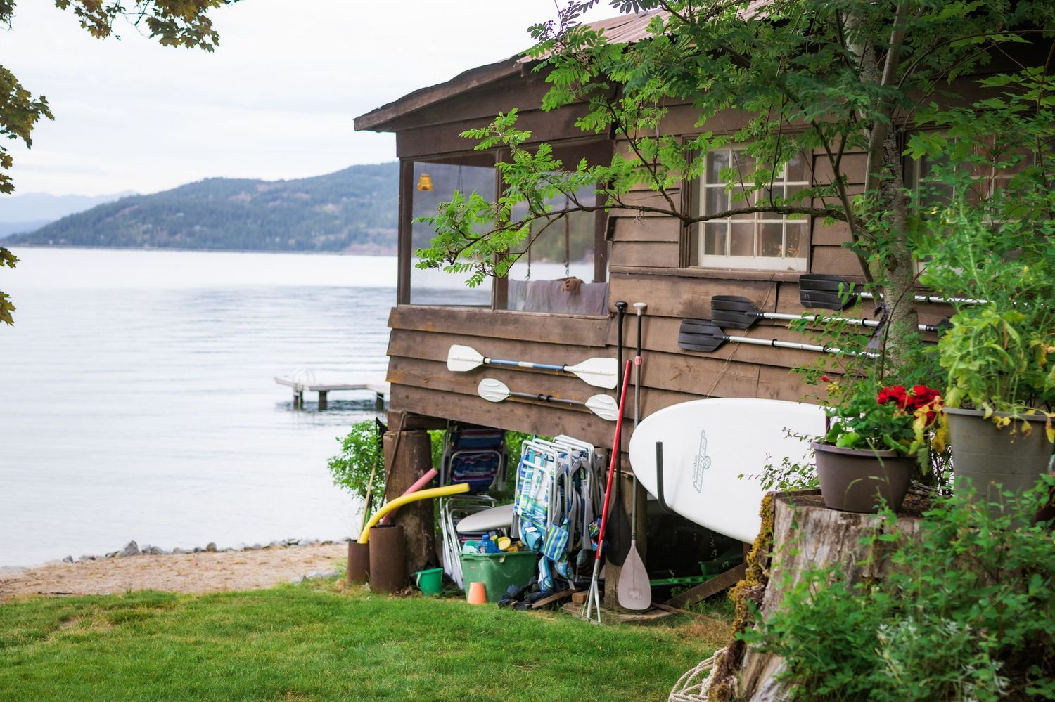 Rustic lakeside boathouse with canoes, paddles, and water toys surrounded by lush greenery.