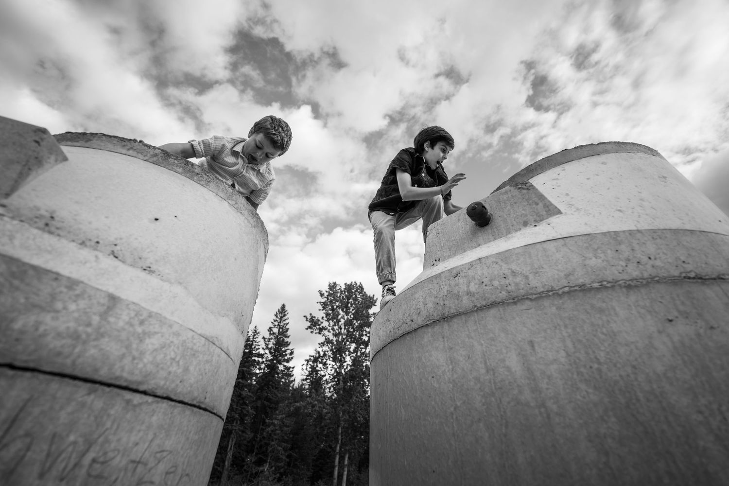 Two children climbing large concrete cylinders outdoors, with trees and cloudy sky in black and white photo.