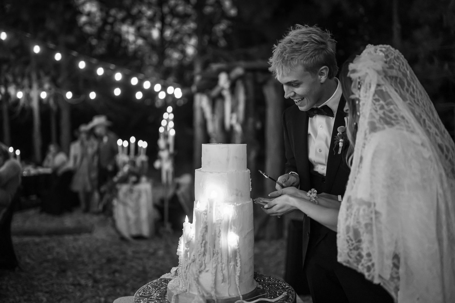 Black and white photo of couple cutting tiered wedding cake outdoors by candlelight.