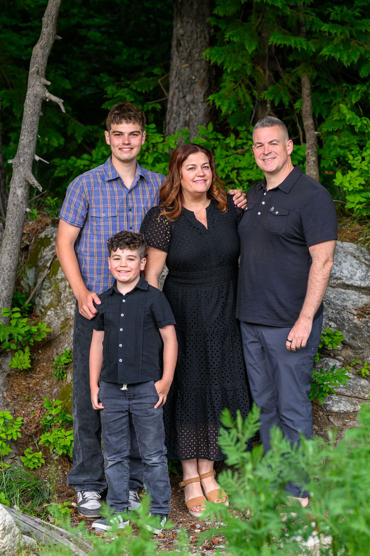 Family of four smiling outdoors among green trees and rocks, posing for a portrait in a lush forest setting.