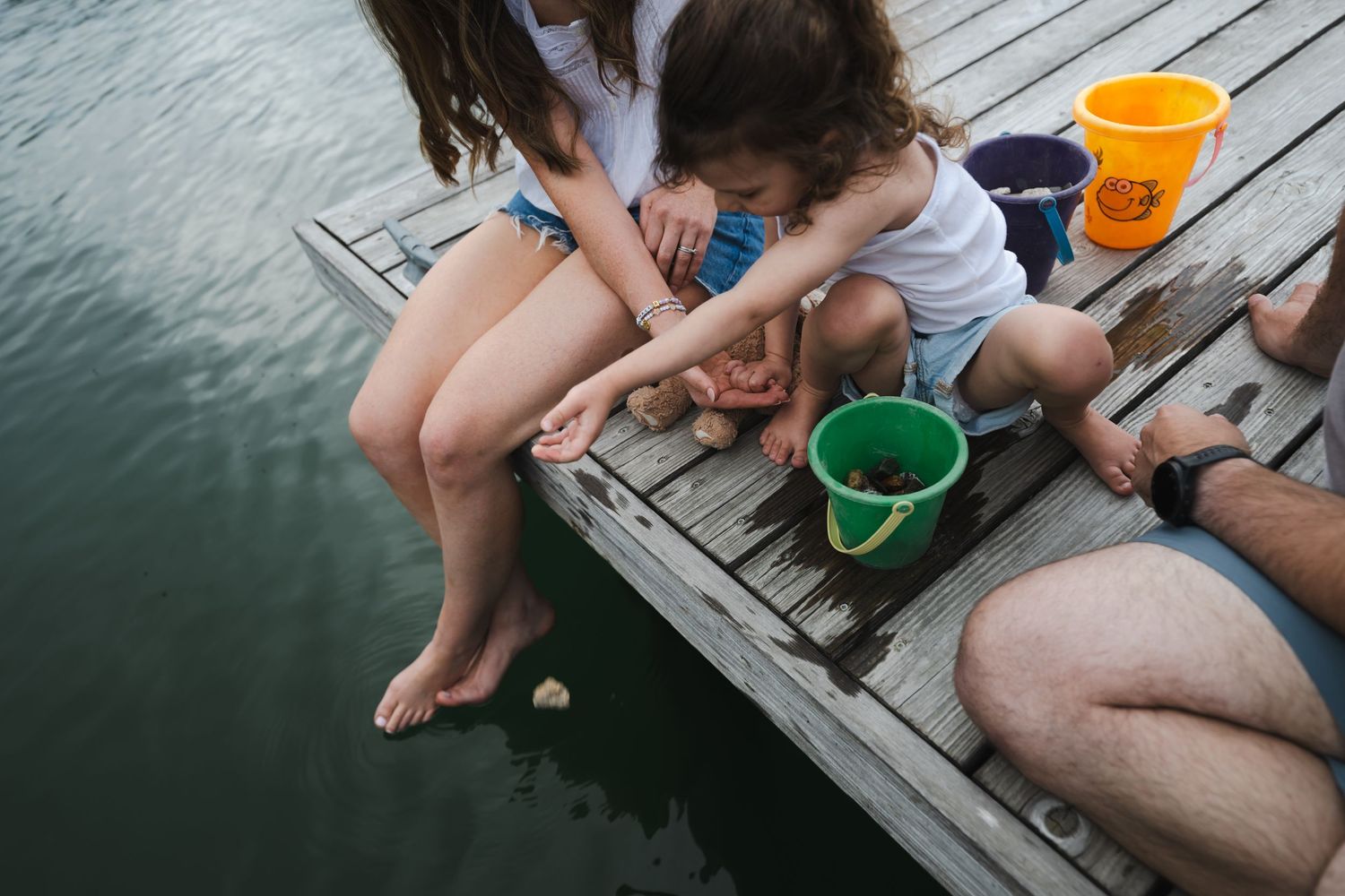 Children and adults sitting on a wooden dock by the water, playing with colorful buckets on a summer day.