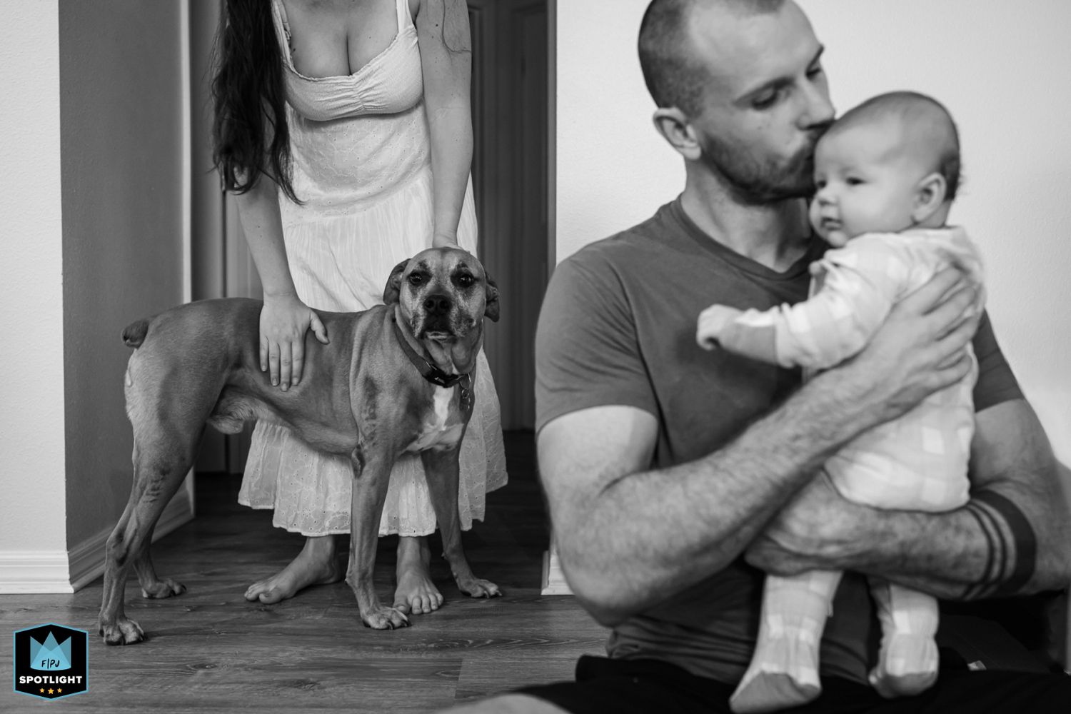 Black and white family portrait with a boxer dog standing beside adults and a baby in a casual indoor setting.