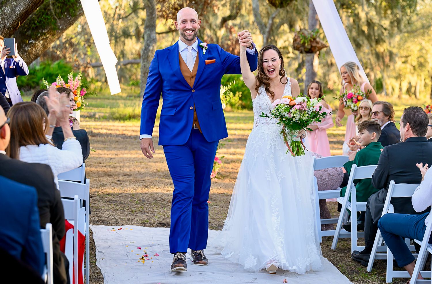 Wedding ceremony outdoors with guests seated as couple exits down aisle draped in white decor.