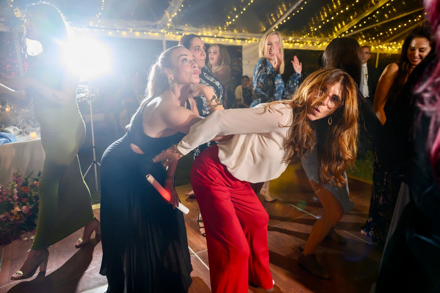 People dancing energetically at a party under string lights in what appears to be a tent or outdoor venue.