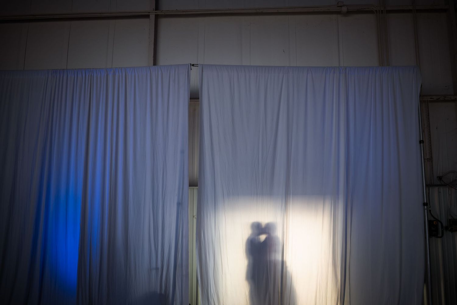 Romantic silhouette of bride and groom kissing behind sheer white curtain with blue ambient lighting.