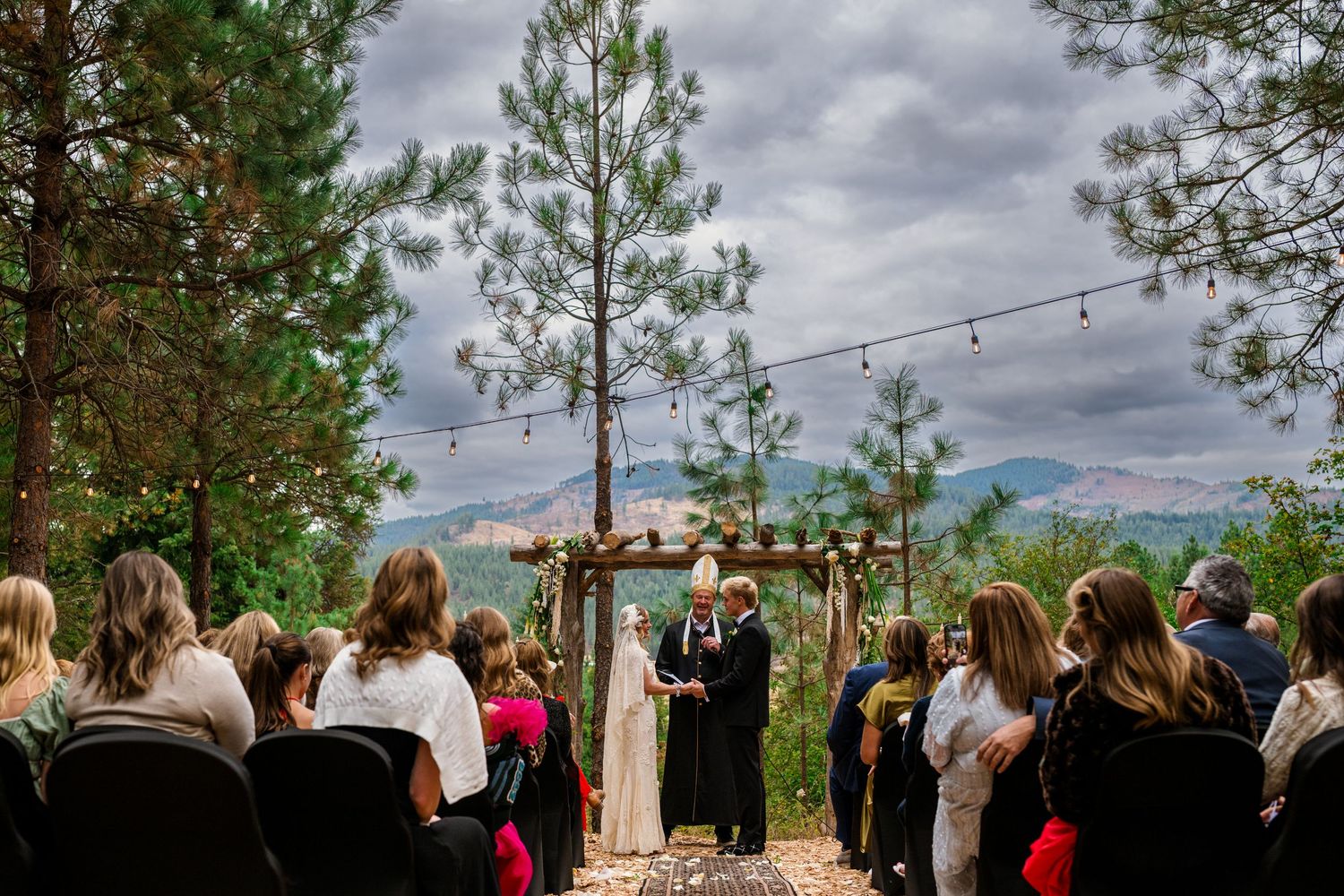 Outdoor wedding ceremony in a pine forest with mountain views, string lights, and guests seated before a wooden arch.