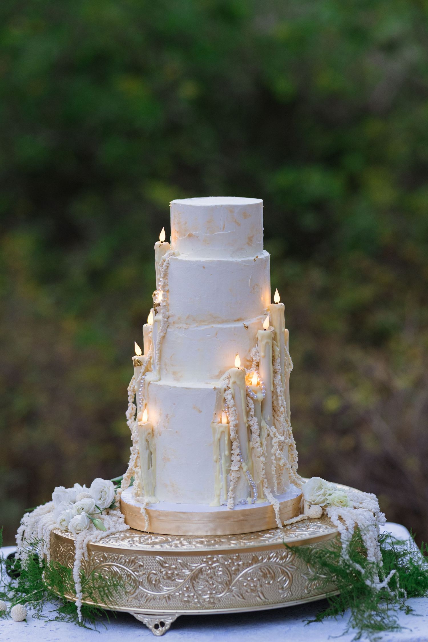 Elegant four-tier white wedding cake adorned with lit candles and gold accents on an ornate cake stand outdoors.