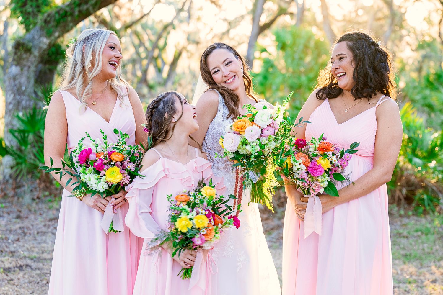 Bridesmaids in flowing pink dresses share laughter while holding colorful spring bouquets in an outdoor garden setting.