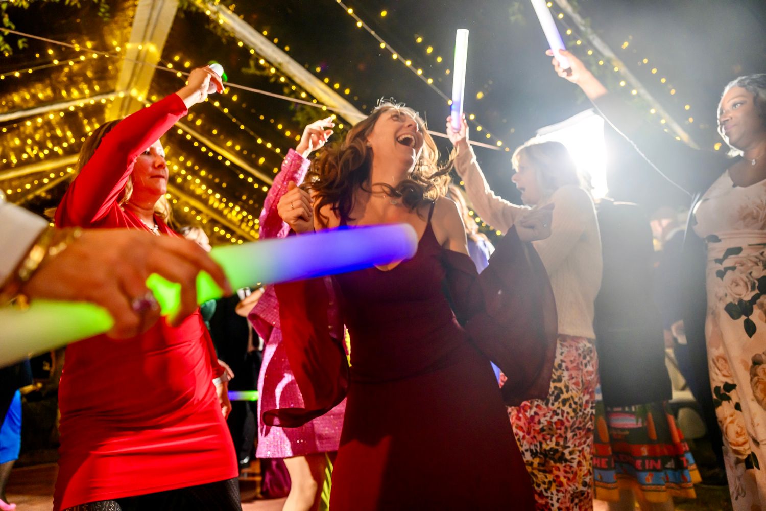 Party guests dance with light-up foam sticks under twinkling string lights at a festive celebration.