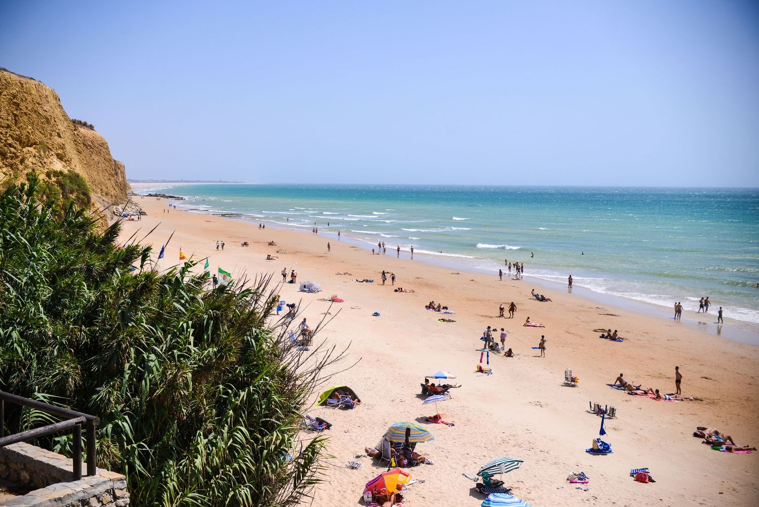 Aerial panoramic view of a sandy beach with scattered beachgoers and umbrellas along turquoise Mediterranean waters.