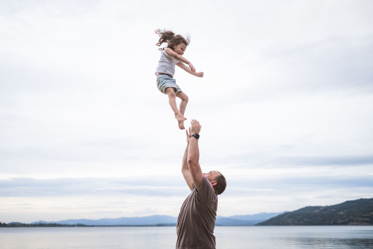 Father joyfully tosses young daughter high into the air near a scenic lakeside with mountains in the background.