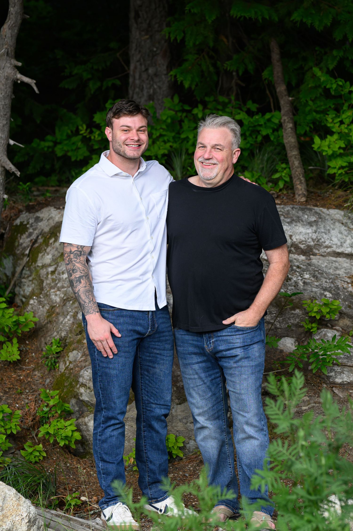 Two smiling men, one tattooed in white shirt and one in black tee, pose together outdoors among lush green trees.