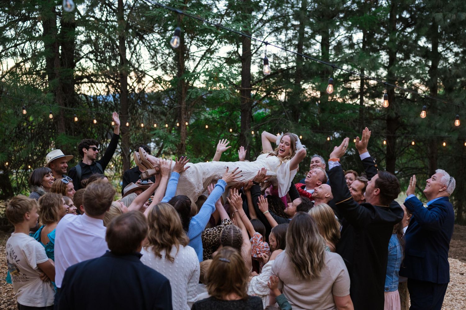 Wedding guests joyfully lifting the bride above their heads during an outdoor forest reception celebration.