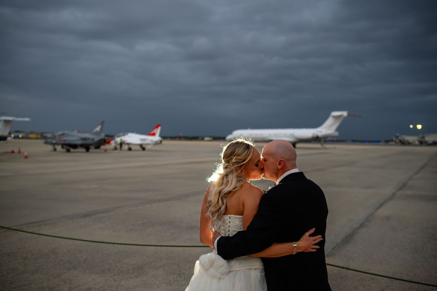 Bride and groom embrace on airport tarmac with private jets and stormy sky backdrop for dramatic wedding photo.