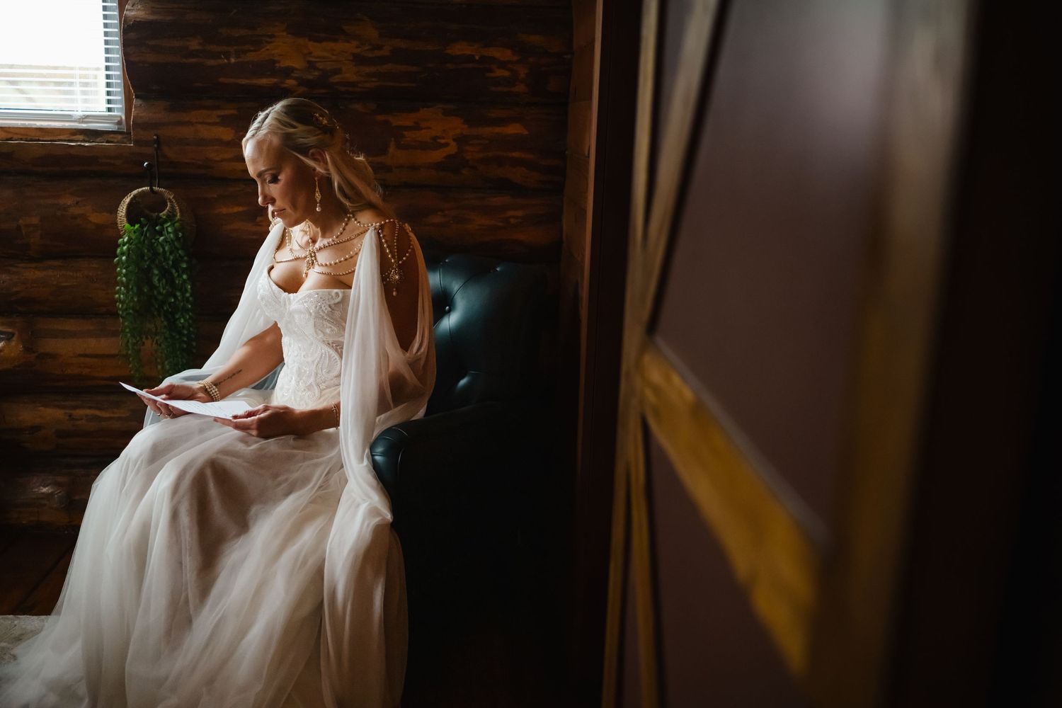 Bride in white tulle gown reading a letter while seated on a leather chair in a rustic log cabin setting.