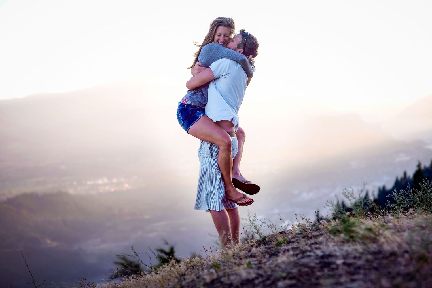 A romantic couple embraces on a hilltop at sunset, man lifting woman in a loving hug with scenic valley below.