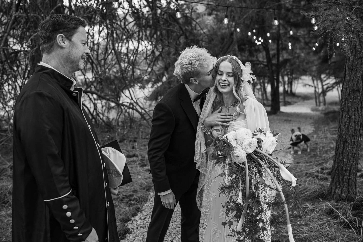 Black and white photo of a bride and groom sharing a moment with their officiant during an outdoor wedding ceremony.