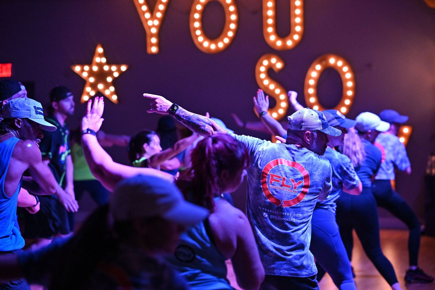 Dancers move in unison under purple lighting with illuminated YOU sign in background.