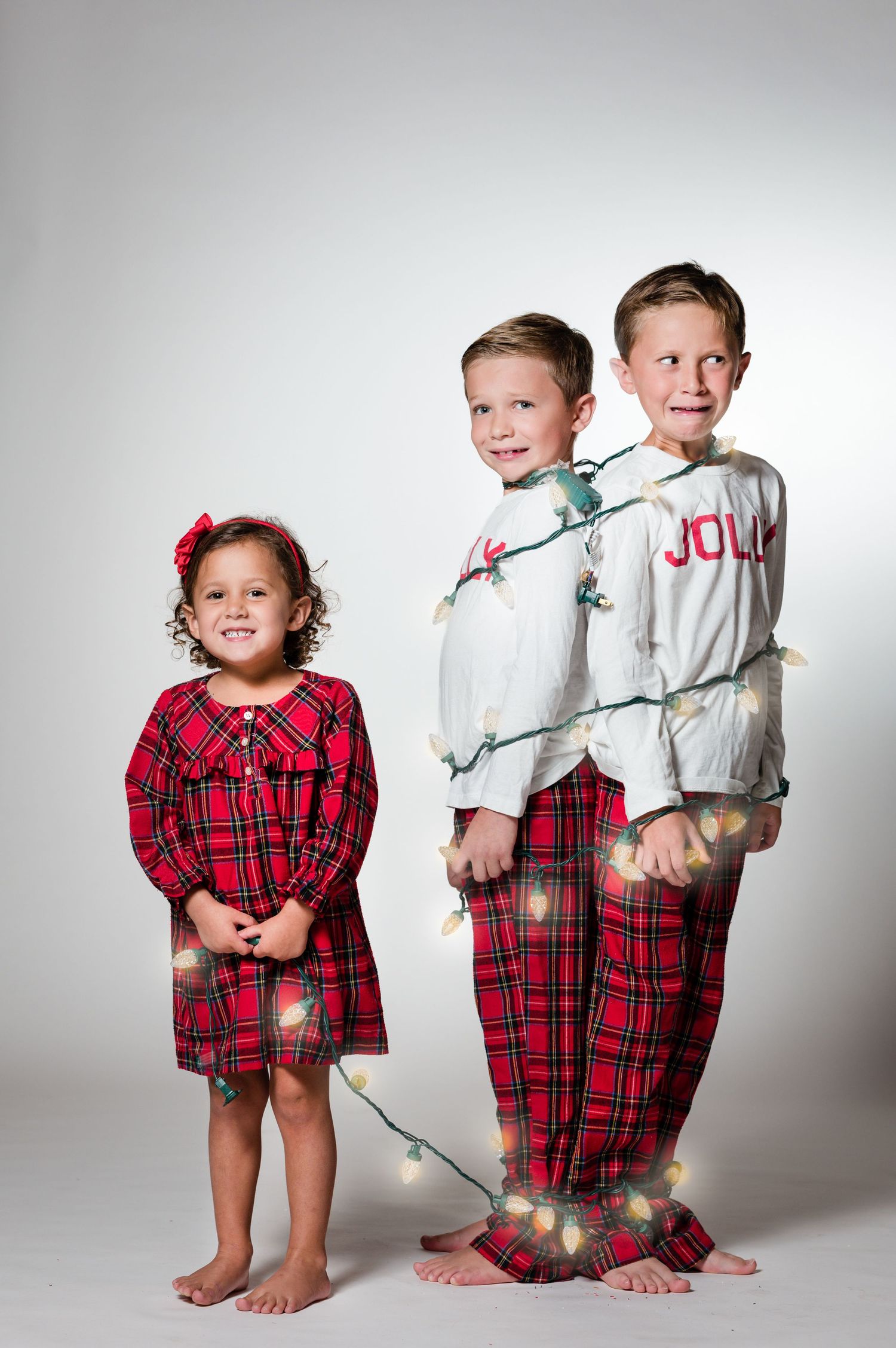 Three children in red plaid Christmas pajamas wrapped in holiday lights pose for a festive holiday photo.