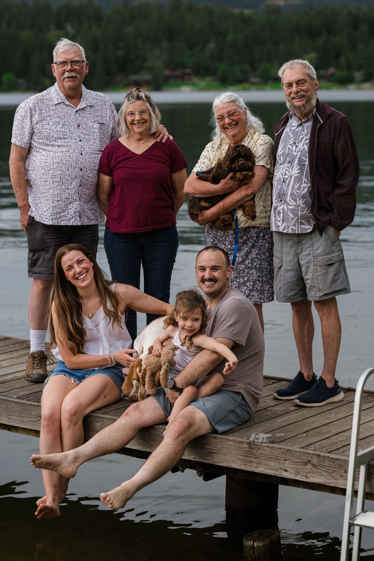 Multi-generational family smiling together on a wooden lake dock with dogs and mountains in the background.