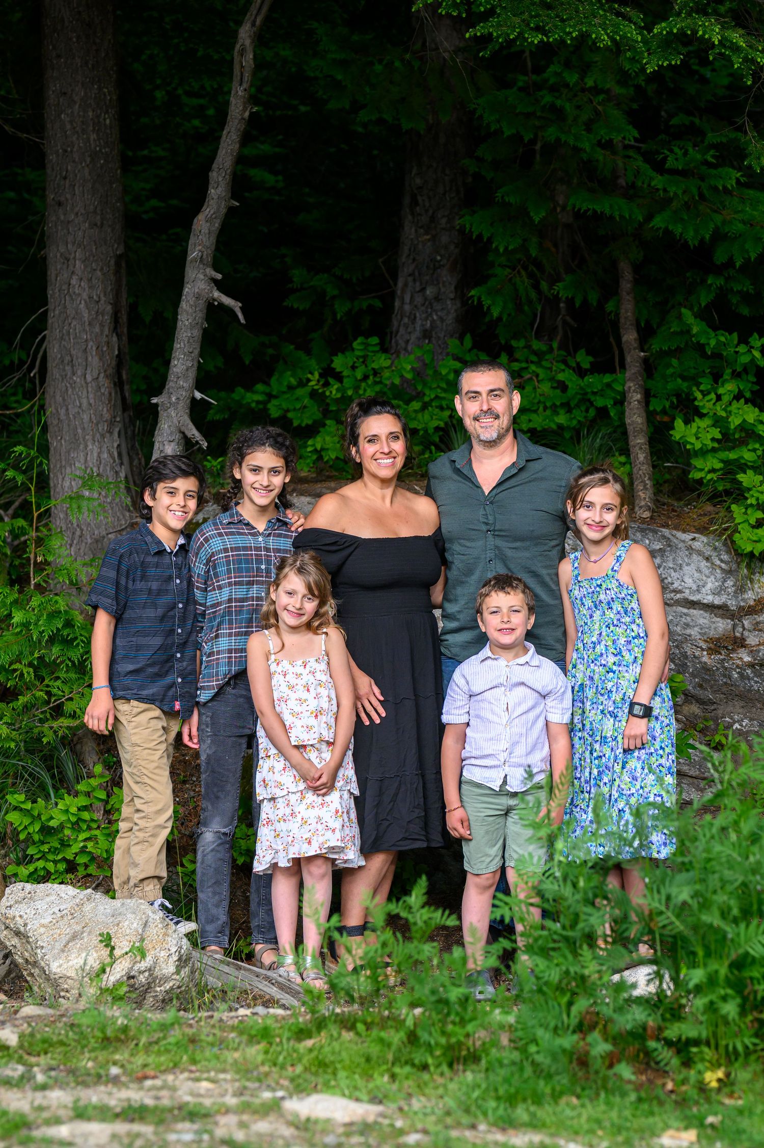 Family of seven smiling together outdoors in a lush forest setting during summer.