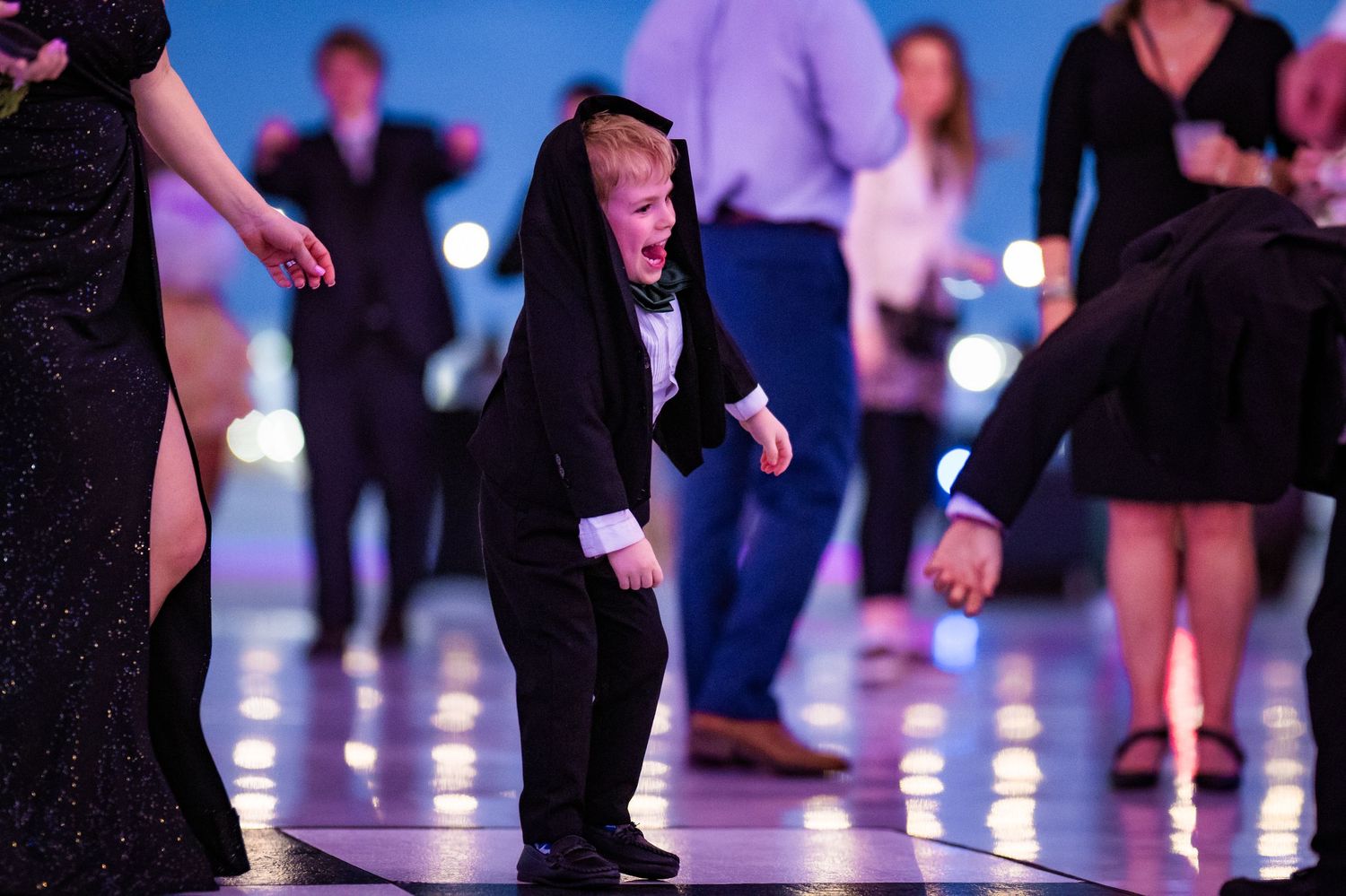 Adorable young boy in a tuxedo dancing energetically on the dance floor at a formal event.