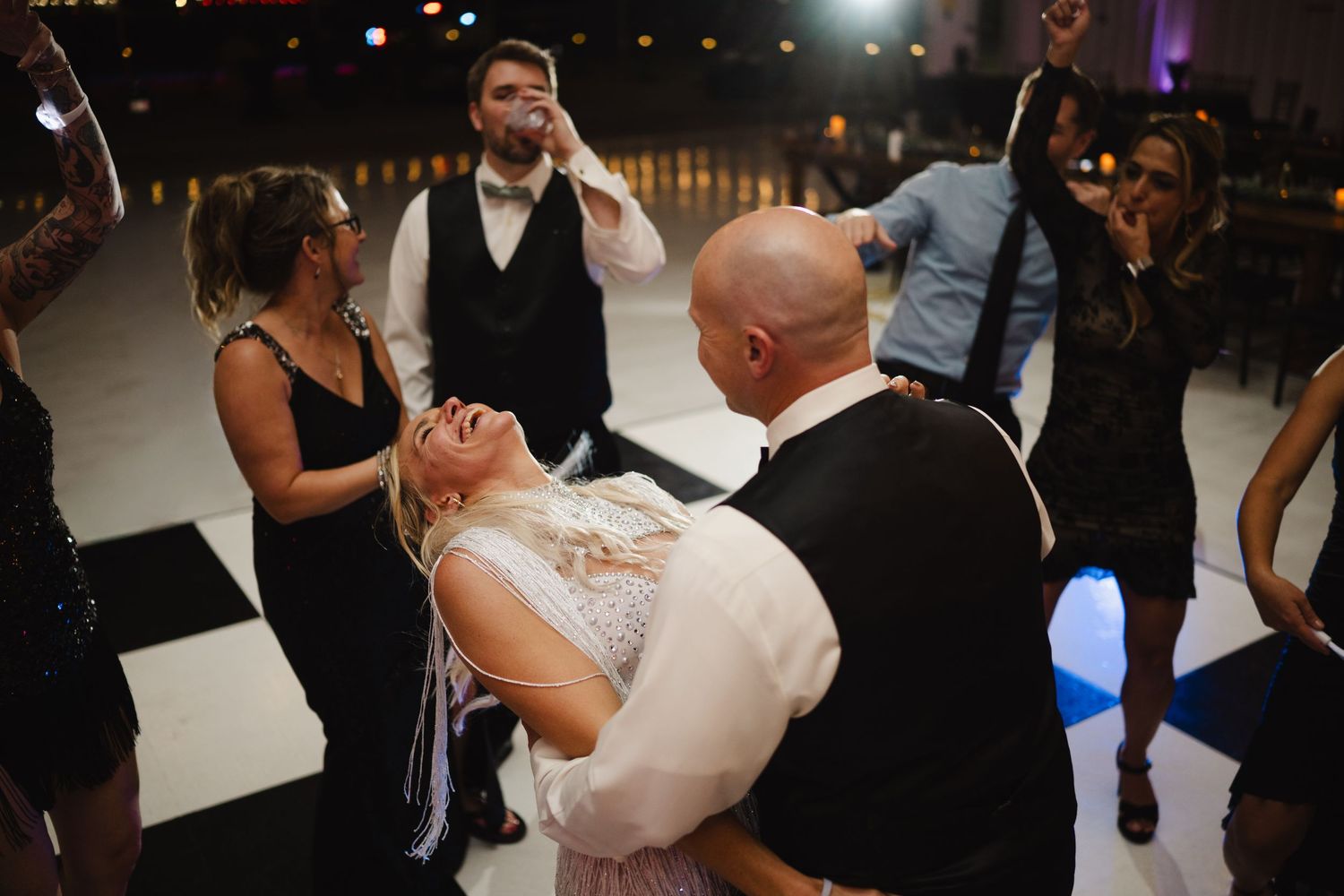 Groom dipping bride on checkered dance floor surrounded by celebrating wedding guests.