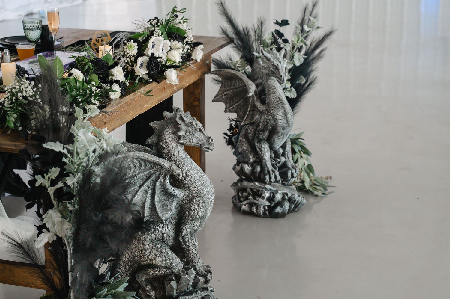Stone dragon statues decorate a rustic wooden sweetheart table with white roses and greenery at a fantasy-themed wedding reception.