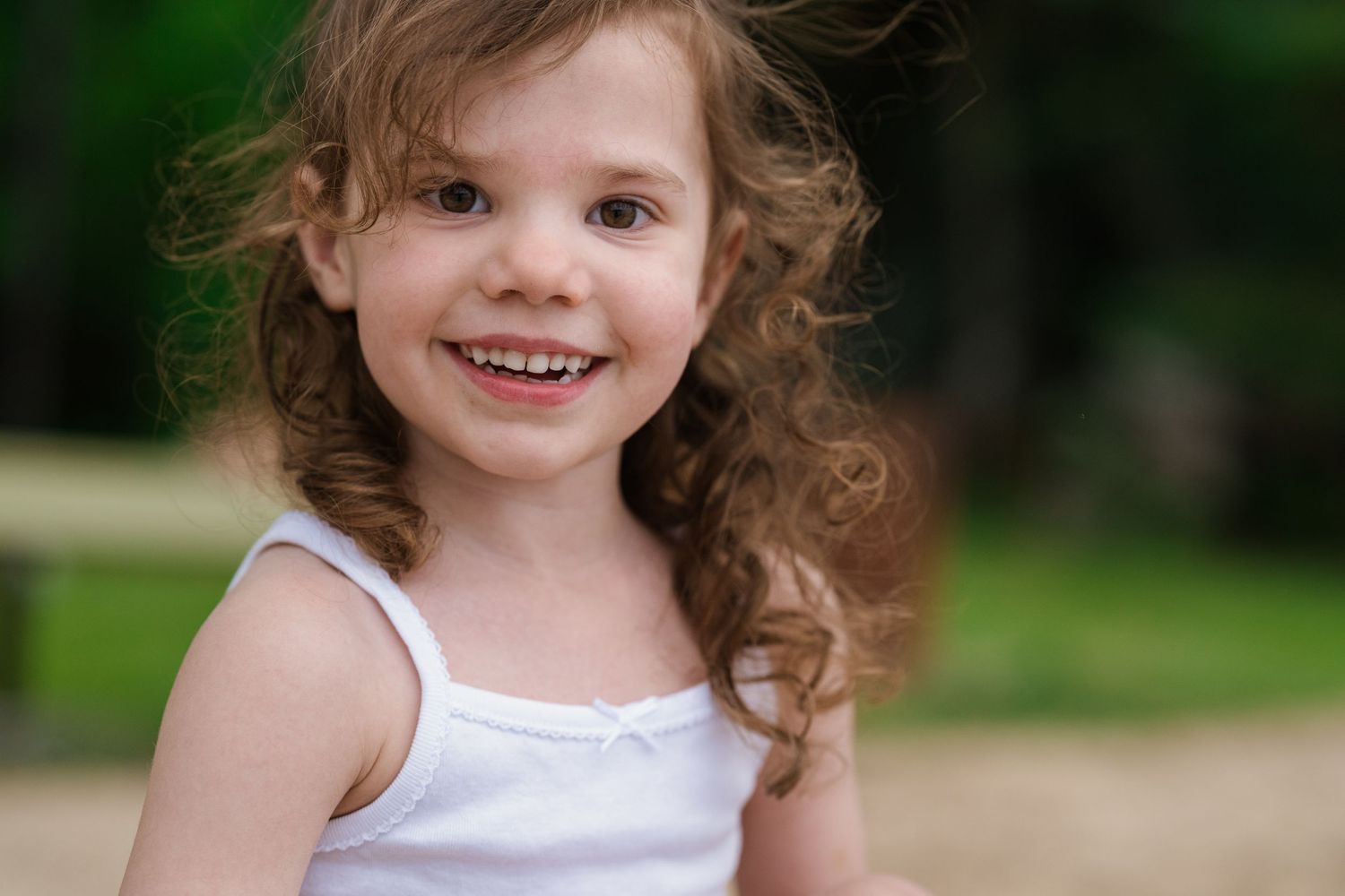 Adorable smiling little girl with curly brown hair wearing a white tank top outdoors.