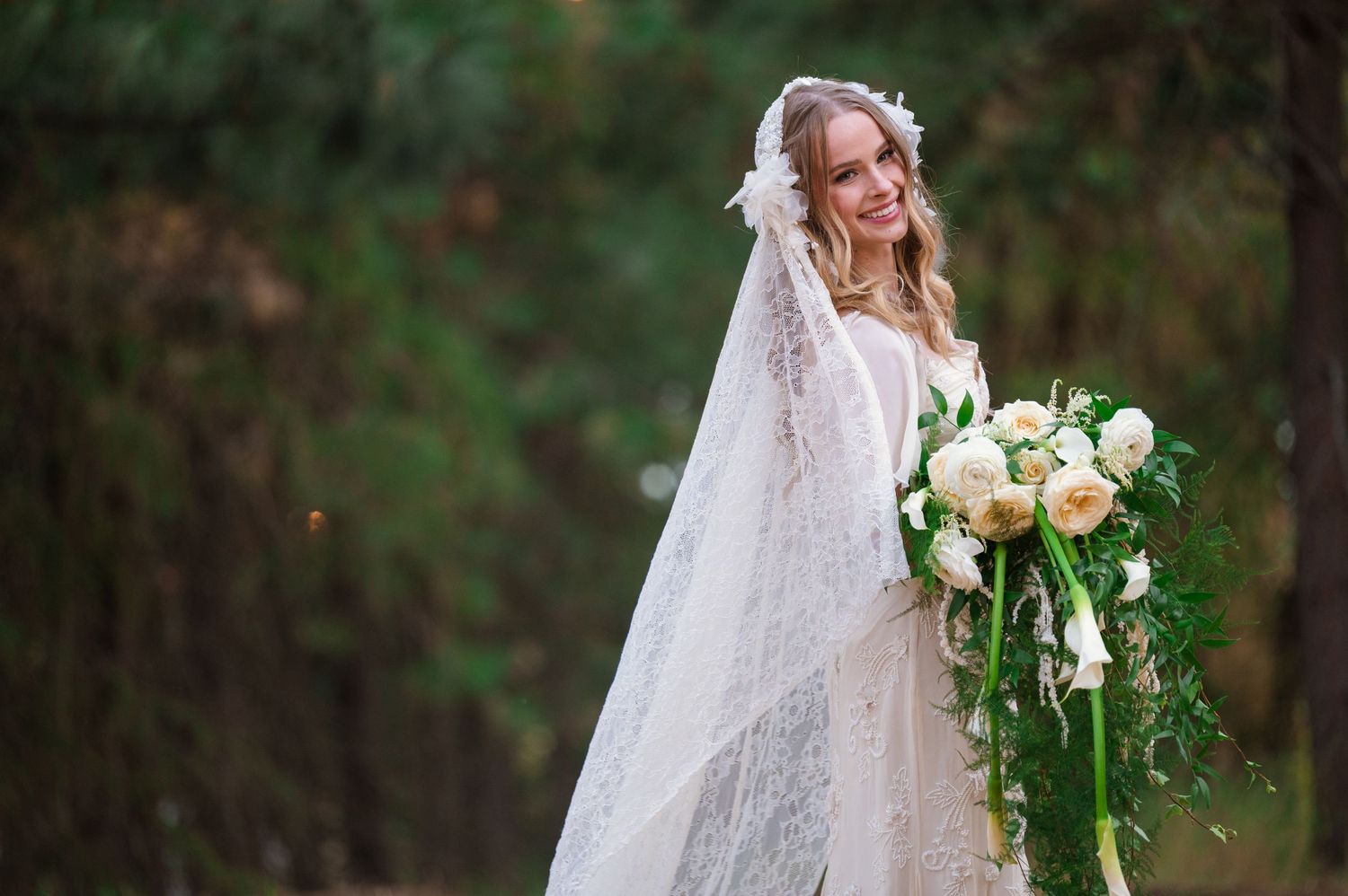 Smiling bride in lace gown and veil holds large white and blush rose bouquet in forest setting.