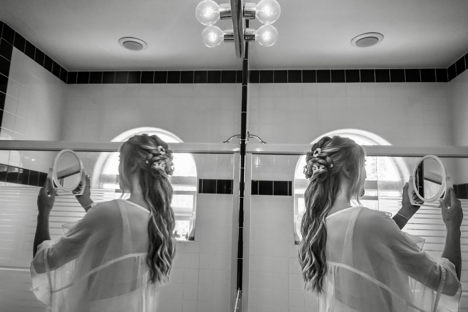 Black and white reflection in bathroom mirror shows long-haired figure in white robe reaching up to adjust their hair.