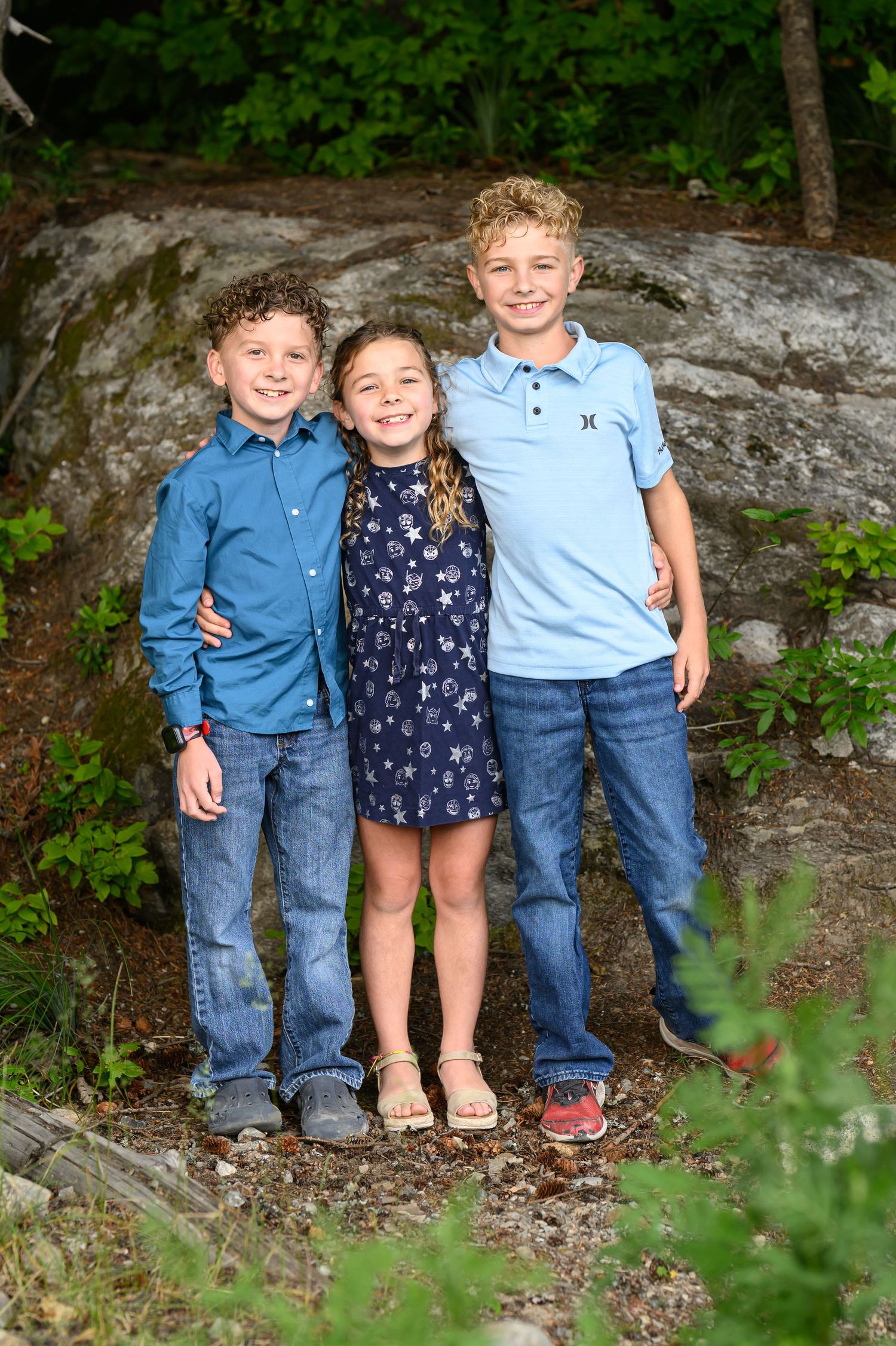 Three smiling children pose together outdoors near a large rock, wearing blue outfits and jeans for a family portrait.