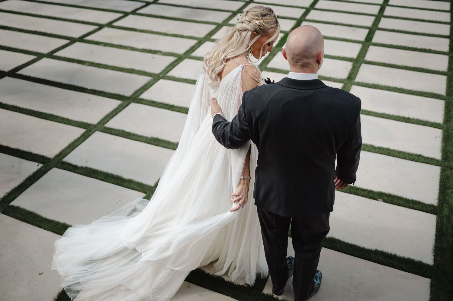 Aerial view of bride in flowing white gown and groom walking together on geometric paved path.