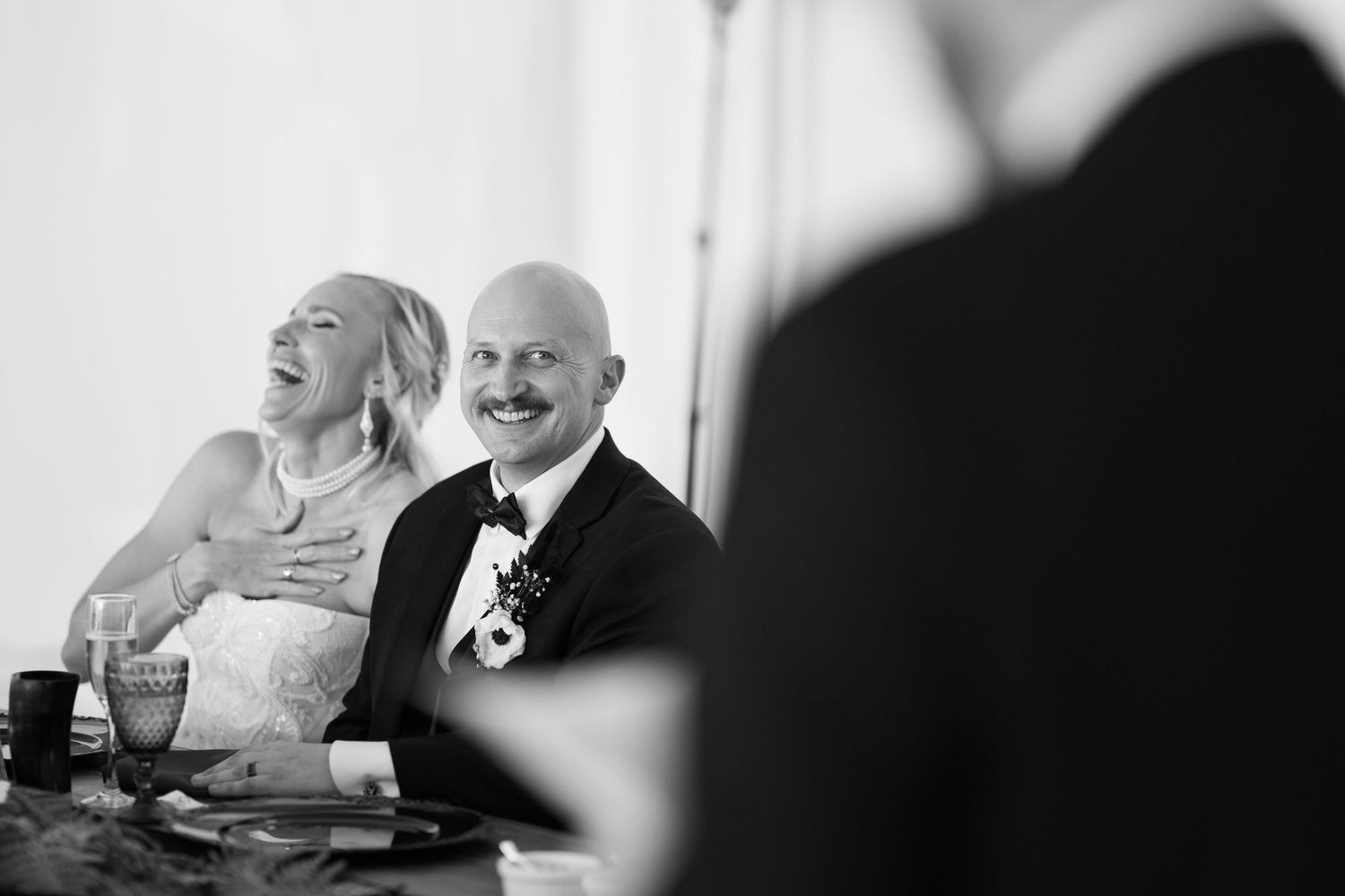 Bride and groom laugh joyfully during wedding reception speeches in black and white photo.