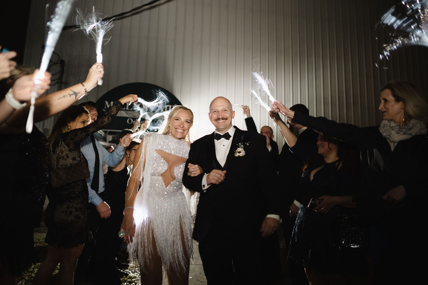 Bride and groom exit wedding reception through sparkler tunnel surrounded by cheering guests at night.