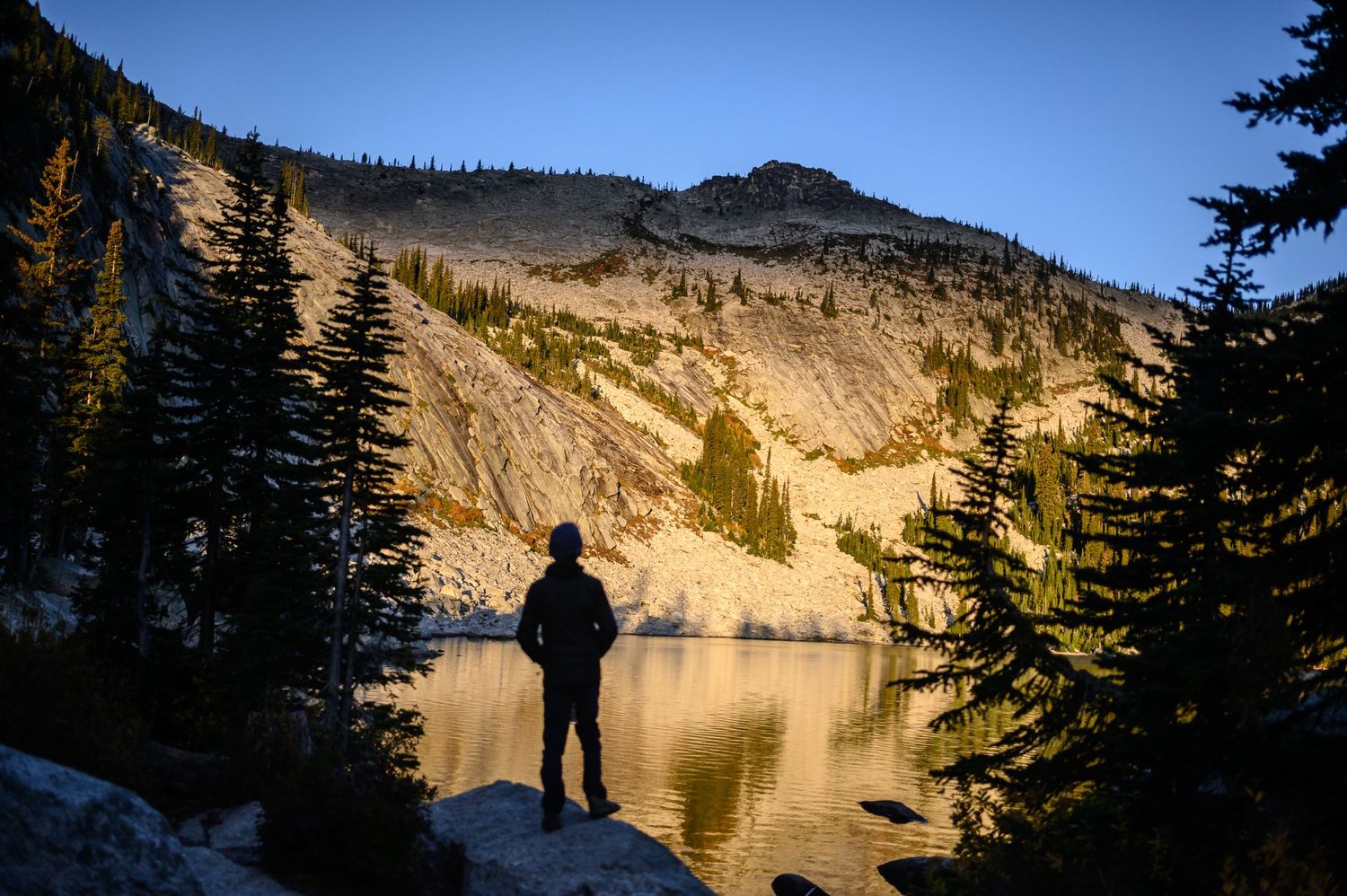 A scenic mountain lake surrounded by evergreen trees reflects golden sunlight on calm waters at sunset.