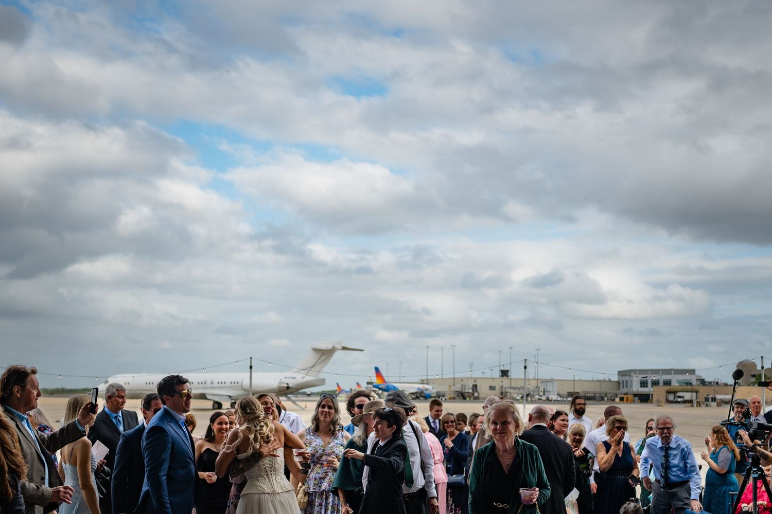 Crowd of wedding guests gathered on airport tarmac with aircraft and cloudy sky in background.